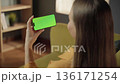 Young woman relaxing at home holding a horizontal smartphone with a green chromakey screen, providing space to add custom content or video calls in living room 136171254