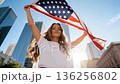 Camera moving to low angle view. Proud serious young woman standing on street holding usa flag under blue sky looking at camera. 136256802
