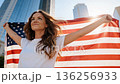 Camera moving to low angle view. Proud serious young woman standing on street holding usa flag under blue sky looking at camera. 136256933