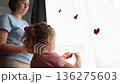 Bottom view of happy little Caucasian girl and her mother decorate the curtains on the window with red paper hearts. Concept of Mother's Day and Valentine's Day. 136275603
