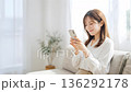 リビングでスマホを操作する若い女性。背景は白いカーテンのある部屋 136292178