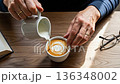 Senior man creating latte art by pouring steamed milk into coffee cup with heart pattern on wooden table with reading glasses and book nearby 136348002