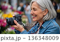 Senior European woman photographer taking photos with professional DSLR camera at colorful outdoor flower market wearing denim jacket 136348009