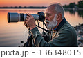 Senior European photographer with gray beard capturing sunset wildlife photos by lake using professional telephoto camera lens on tripod 136348015