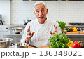 Senior European Chef in White Uniform Explaining Cooking Techniques with Hand Gestures in Modern Professional Kitchen with Fresh Vegetables 136348021