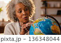 Senior African American woman with curly gray hair examining world globe while planning travel destinations in cozy home study room 136348044