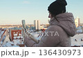 Woman using smartphone outdoors in winter while standing on balcony with city view 136430973