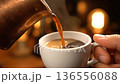 A hand carefully pours steaming hot coffee into a small white cup, creating a cozy atmosphere. 136556088