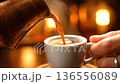 Fresh hot coffee is carefully poured from a copper pot into a small white cup. 136556089