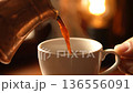 Close up of hand pouring fresh, dark coffee into white ceramic cup, warm background. 136556091