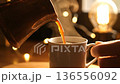 Hot coffee is poured into a white ceramic mug, creating a cozy and warm atmosphere. 136556092