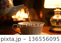 A warm stream of hot tea pours from a teapot into a cup. Cozy evening by the fireplace. 136556095