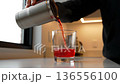 A hand pouring a refreshing red beverage from a travel tumbler into a clear glass, indoors. 136556100