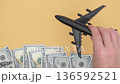 Hand holding black airplane toy over stacks of cash on yellow background, showcasing imaginative travel planning, camera follows action upward 136592521