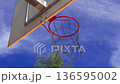 A stunning view of a basketball hoop glistens against the backdrop of bright blue skies. Nearby trees add to the lively atmosphere of the bustling park where players gather. 136595002
