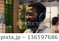 African american man in good mood listens to music on headset, working with laptop and combining stress relief with focused business analysis. Multitasking with audio and tasks in office. 136597686