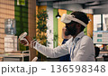 Black professional using VR glasses in corporate office environment. High-tech virtual reality applications display charts and performance metrics, combining metaverse simulation and data analysis. 136598348