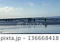 Group of surfers in wetsuits wading into the sea with boards, facing breaking waves on a sandy beach. Wet shoreline reflects sky, ideal for travel, sport, and outdoor lifestyle. 136668418