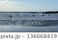 Group of surfers in wetsuits wading into the sea with boards, facing breaking waves on a sandy beach. Wet shoreline reflects sky, ideal for travel, sport, and outdoor lifestyle. 136668419