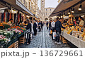 bustling cobblestone street market flowers produce. vendors display colorful bouquets, fresh fruit, cheeses, artisanal breads tourists and locals browse stalls under canvas awnings 136729661