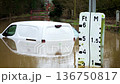 Flood warning sign measuring water level while van remaining submerged on rural road. Brown river water rising and surpassing gauge markings during severe storm. Climate crisis worsening and causing 136750817