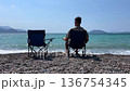 Back view of a man sitting alone on a pebble beach beside an empty chair facing the sea. Concept of loneliness, solitude, reflection and peaceful summer vacation. 136754345