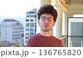 Young man with glasses and curly hair standing on the balcony of his city apartment. He starts talking and then smiles at the camera, exuding confidence and a positive urban lifestyle 136765820