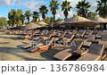 Rows of empty wooden sun loungers with straw umbrellas line a peaceful, sandy beach at a luxury seaside resort. Calm turquoise water and lush green hills under a bright morning sky. High quality 4k 136786984