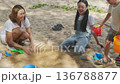 Family beach vacation, happy children play sand with colorful toys, young woman watches kids, enjoying summer travel trip outdoors, creating wonderful memories with engaging fun activity. 136788877