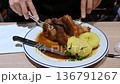 Munich, germany, december 27, 2025. Man's hands using a fork and knife to cut a large piece of roasted pork knuckle. Delicious traditional german schweinshaxe meal served on a plate 136791267