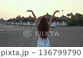 Woman with raised arms on sandy beach at sunset facing resort cabanas 136799790