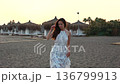 Woman walking on sandy beach at sunset by thatched umbrellas and closed sunbeds 136799913