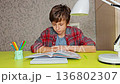 A boy is sitting at a table with a laptop and a book. He is focused on his work and he is in a serious mood. A schoolboy boy learns lessons at the table 136802307