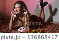 Portrait photo of happy smiling woman with bright makeup holding telephone receiver. Excited retro style girl. Beautiful model in red dress holds pink telephone receiver in hand and poses for photo 136868417