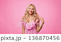 A woman with light skin and long blonde hair smiles confidently while wearing a pink dress. The background is a solid pink color, adding a playful vibe to the scene. A woman with light skin and long blonde hair smiles confidently while wearing a pink dress. The background is a solid pink color, adding a playful vibe to the scene. 136870456