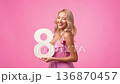 A woman with long blonde hair stands in front of a pink background. She smiles while holding a large white number eight board in her hands. A woman with long blonde hair stands in front of a pink background. She smiles while holding a large white number eight board in her hands. 136870457