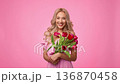 A woman with light skin stands smiling while holding a bouquet of vibrant flowers. The background is a soft pink color. She appears joyful and relaxed in this moment. A woman with light skin stands smiling while holding a bouquet of vibrant flowers. The background is a soft pink color. She appears joyful and relaxed in this moment. 136870458
