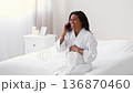 A woman with dark skin sits on a bed in a well-lit room. She is wearing a bathrobe and talking on the phone with a concerned expression while holding her belly. A woman with dark skin sits on a bed in a well-lit room. She is wearing a bathrobe and talking on the phone with a concerned expression while holding her belly. 136870460