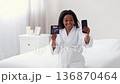 A pregnant woman with dark skin smiles while holding an ultrasound image and using her smartphone. She is sitting on a bed in a bright, cozy room. A pregnant woman with dark skin smiles while holding an ultrasound image and using her smartphone. She is sitting on a bed in a bright, cozy room. 136870464