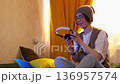 Happy young brunette in glasses wearing headphones sits on bed talking on laptop during video call, concept of internet meeting and distance conversation. Mid shot. 136957574
