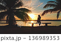 Woman sits peacefully on a bench under palm trees at sunset by the ocean, enjoying the view. The sky is painted in orange and pink hues, creating a serene atmosphere in a tropical paradise 136989353
