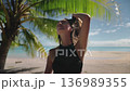 Young woman relaxes under a palm tree on a tropical beach, soaking up the sun and enjoying the turquoise ocean. She embodies a carefree, happy lifestyle in a paradise-like setting 136989355