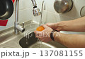 Male hands wash fresh orange under clean water flowing from faucet above sink on home kitchen, close-up. Person removes bacteria, pesticides, rinses fruit before consumption, handheld shot. 137081155