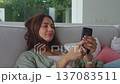 Mobile phone woman smiles while checking smartphone on sofa at home. Mobile phone lifestyle reflects relaxed leisure comfort digital connection calm mood and modern indoor living. 137083511
