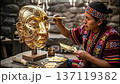 In a workshop, a skilled Inca craftsman works on a copper mask. He slowly applies thin gold leaf with a soft brush. The process shows his attention to detail and the care in each movement. 137119382