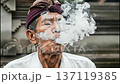 A man in Bali slowly exhales a plume of thick white smoke from a kretek cigarette. He sits outdoors near a temple. The smoke curls upwards into the air, creating a simple moment. 137119385