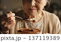 An elderly woman gently sprinkles cocoa powder over a Tiramisu. Her hands show care and skill as she adds the final touch. This takes place in a warm kitchen in the afternoon. 137119389