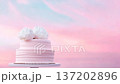 A large two-tier cake stands on a table outside during sunset. The cake has a light pink color and is topped with white flowers. The sky shows shades of pink and blue 137202896