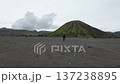 Woman walks alone across black volcanic sand toward lush Mount Batok and smoking Mount Bromo in Indonesia. Scenic travel moment filled with solitude, nature, and adventure. 137238895