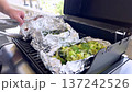 A wide view of a backyard grill in use with multiple foil packets filled with vegetables. The suburban setting and visible hands evoke a homey, relaxed cooking moment. 137242526